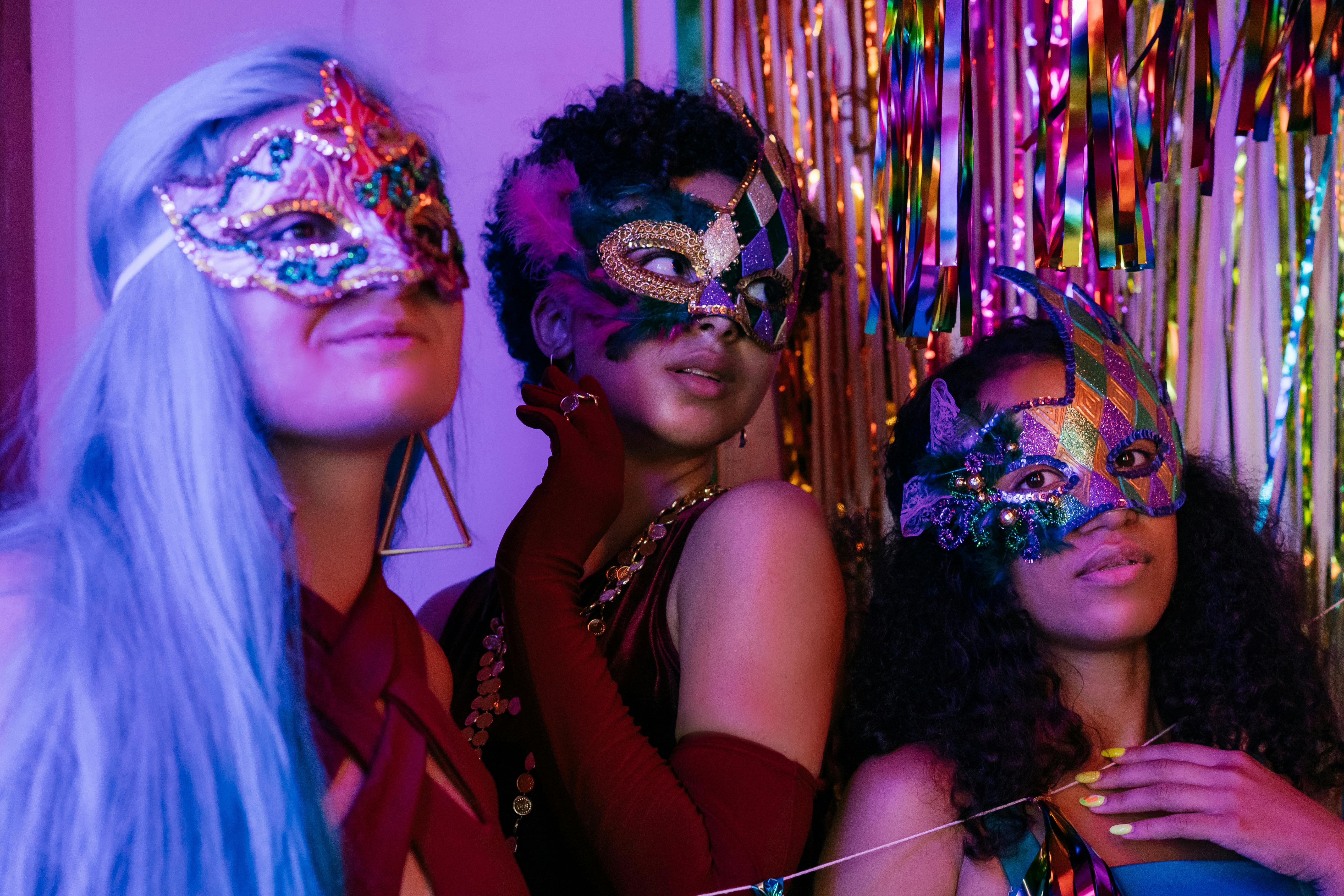 Three people wearing colorful masquerade masks, smiling under festive lights.