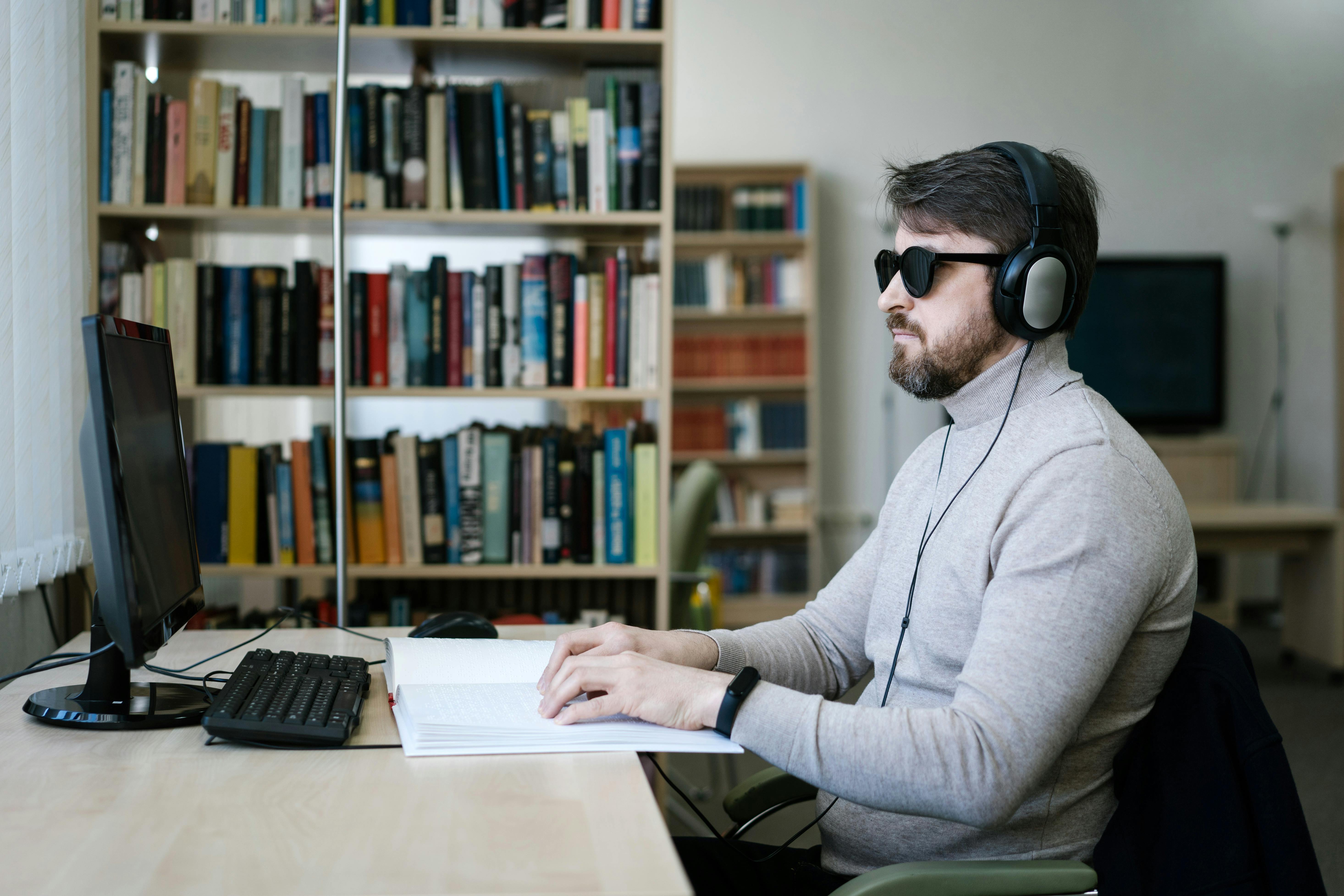 Man using a computer and braille book, wearing headphones and sunglasses.